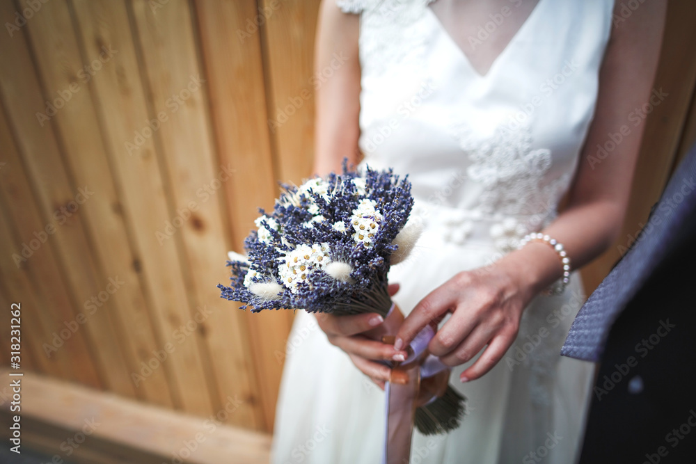 Selective focus unusual bouquet of wild flowers chamomile, lavender, ears in the hands of an crop bride in a white wedding dress. Selective focus unusual bouquet of wild flowers chamomile, lavender, ears in the hands of an crop bride in a white wedding dress.