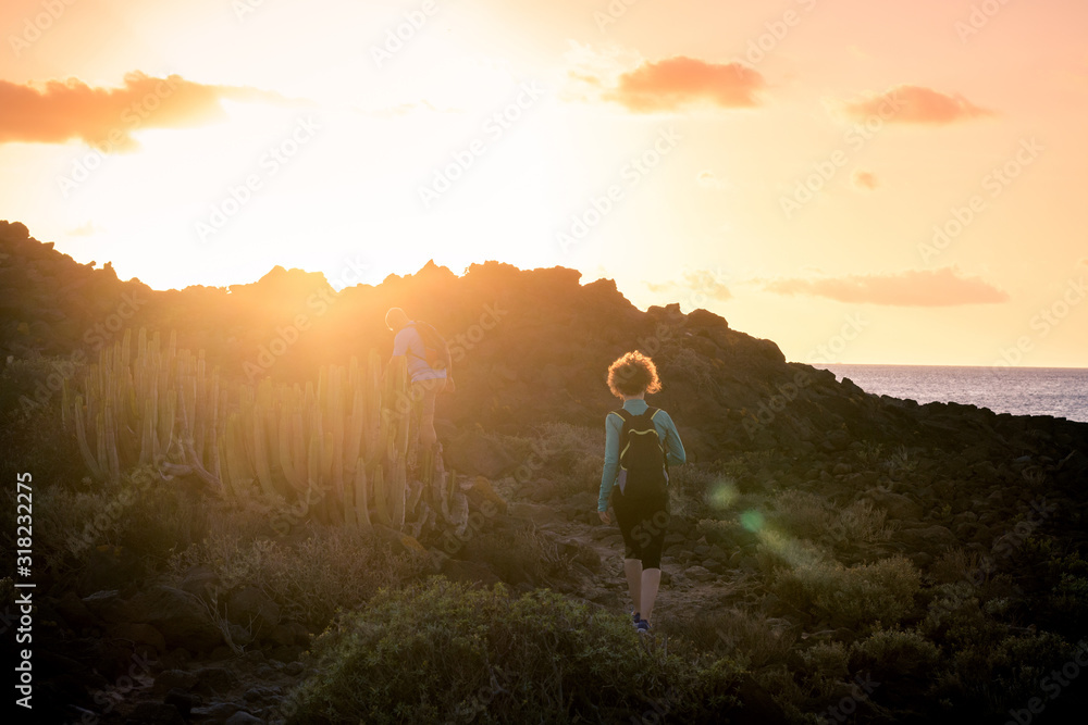 Back view of a couple of sporty trekkers hiking in a volcanic landscape near the ocean in Canary Island. Man and woman with backpack walking together in a holiday evening at sunset. Sport concept.