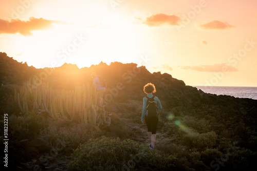 Back view of a couple of sporty trekkers hiking in a volcanic landscape near the ocean in Canary Island. Man and woman with backpack walking together in a holiday evening at sunset. Sport concept.