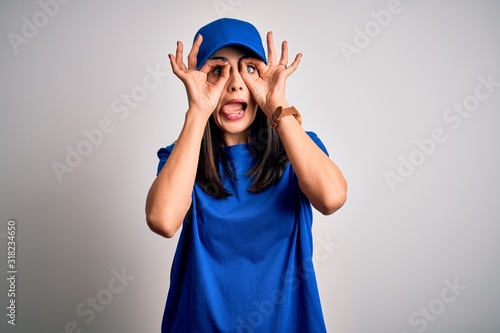 Young delivery woman with blue eyes wearing cap standing over blue background doing ok gesture like binoculars sticking tongue out, eyes looking through fingers. Crazy expression.