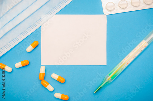Medical mask, orange and white capsules, thermometer, pills on a blue background and a white sheet with copy space. First aid, care during illness.
