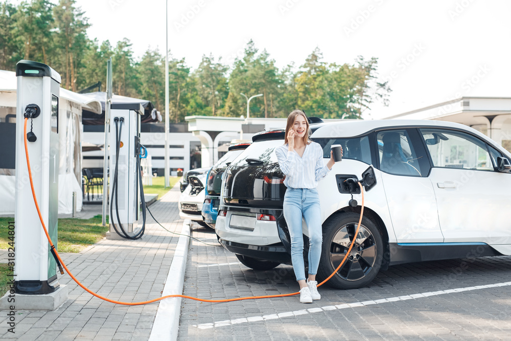 Transportation. Young woman on electric car having stop at charging station leaning on vehicle drinking hot coffee talking on smartphone smiling joyful while charging Transportation. Young woman on electric car having stop at charging station leaning on vehicle drinking hot coffee talking on smartphone smiling joyful while charging