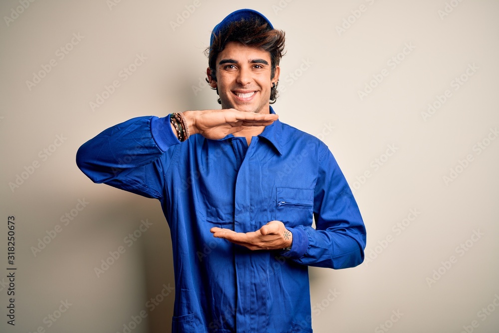 Young mechanic man wearing blue cap and uniform standing over isolated white background gesturing with hands showing big and large size sign, measure symbol. Smiling looking at the camera. Measuring Young mechanic man wearing blue cap and uniform standing over isolated white background gesturing with hands showing big and large size sign, measure symbol. Smiling looking at the camera. Measuring