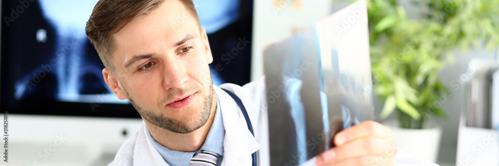 Fototapeta premium Portrait of concentrated man indicating something on x-ray picture with pencil. Physician wearing white uniform and stethoscope carefully examining sciagram. Medicine concept. Blurred background