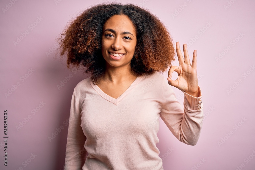 Young african american woman with afro hair wearing casual sweater over pink background smiling positive doing ok sign with hand and fingers. Successful expression. Young african american woman with afro hair wearing casual sweater over pink background smiling positive doing ok sign with hand and fingers. Successful expression.