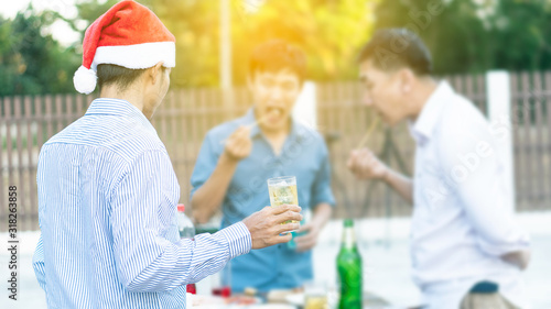 One man holds a glass of beer and two men are eating barbecue behind him and the orange light.
