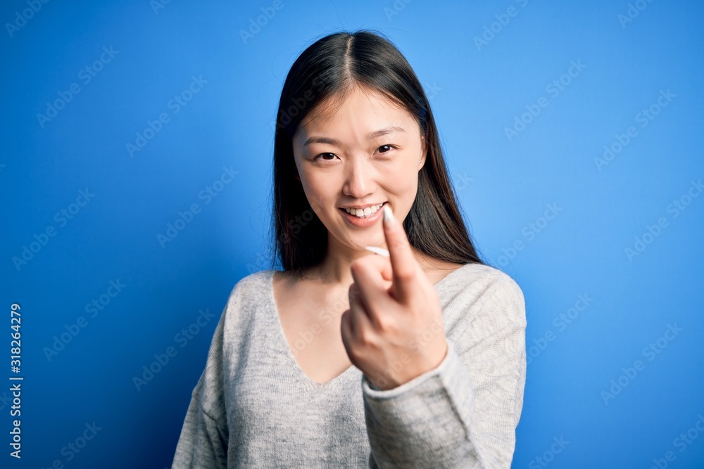 Young beautiful asian woman wearing casual sweater standing over blue isolated background Beckoning come here gesture with hand inviting welcoming happy and smiling Young beautiful asian woman wearing casual sweater standing over blue isolated background Beckoning come here gesture with hand inviting welcoming happy and smiling