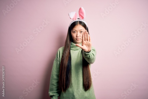Photography Young asian woman wearing cute easter bunny ears over pink background doing stop sing with palm of the hand