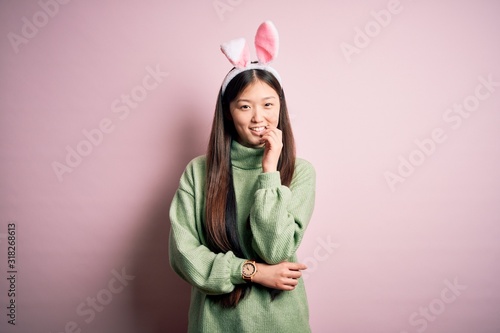 Young asian woman wearing cute easter bunny ears over pink background looking confident at the camera with smile with crossed arms and hand raised on chin. Thinking positive.