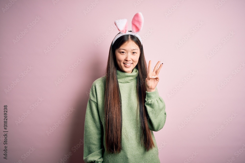 Young asian woman wearing cute easter bunny ears over pink background showing and pointing up with fingers number three while smiling confident and happy. Young asian woman wearing cute easter bunny ears over pink background showing and pointing up with fingers number three while smiling confident and happy.