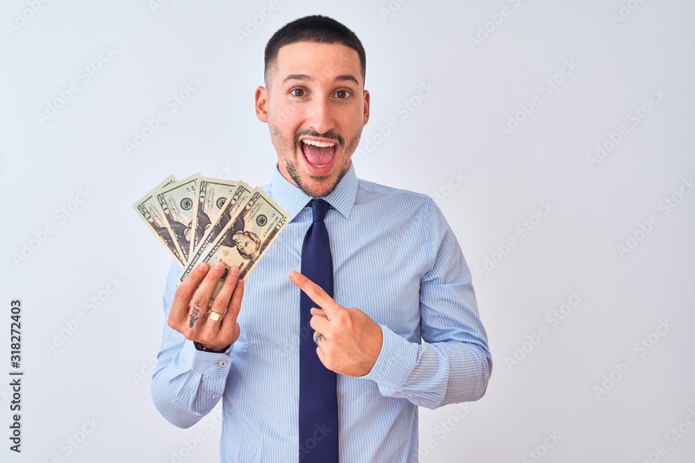 Young handsome business man holding a bunch of bank notes dollars over isolated background very happy pointing with hand and finger Young handsome business man holding a bunch of bank notes dollars over isolated background very happy pointing with hand and finger