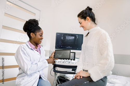 African female doctor gynecologist or obstetrician showing ultrasound photo of baby to young pregnant woman. Ultrasound photo, gynecology concept.