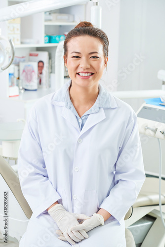 Vertical medium long shot of attractive Asian female dentist sitting in her office looking at camera  smiling