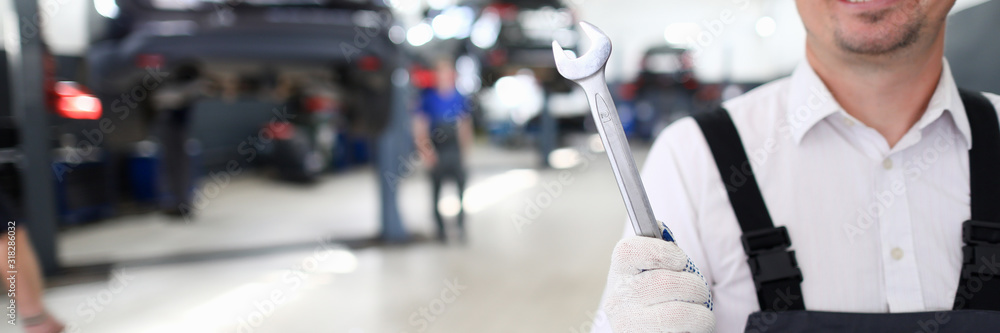 Portrait of cheerful man holding shining metal wrench and standing in modern car maintenance garage with many automobiles. Machinery repairman and service station concept