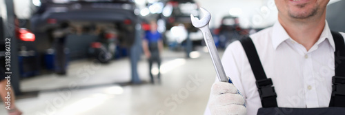 Portrait of cheerful man holding shining metal wrench and standing in modern car maintenance garage with many automobiles. Machinery repairman and service station concept © H_Ko