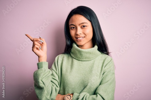 Young beautiful chinese woman wearing turtleneck sweater over isolated pink background with a big smile on face, pointing with hand and finger to the side looking at the camera.