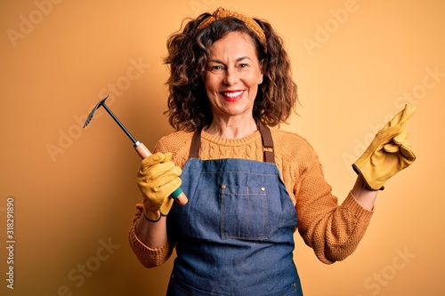 Middle age gardener woman wearing apron holding rake over isolated yellow background very happy pointing with hand and finger to the side