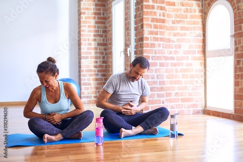 Middle age sporty couple sitting on mat doing stretching yoga exercise at gym with hand on stomach because nausea, painful disease feeling unwell. Ache concept.