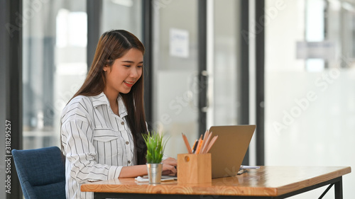 Young business girl in striped shirt typing on laptop and sitting at the wooden working desk with modern office background.