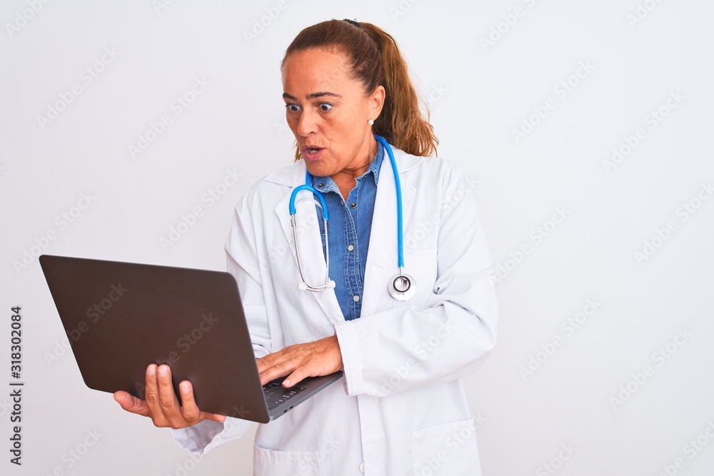 Middle age mature doctor woman holding computer laptop over isolated background scared in shock with a surprise face, afraid and excited with fear expression Middle age mature doctor woman holding computer laptop over isolated background scared in shock with a surprise face, afraid and excited with fear expression