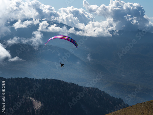 A man on a paraglider flies through the clouds over the forest and mountains.