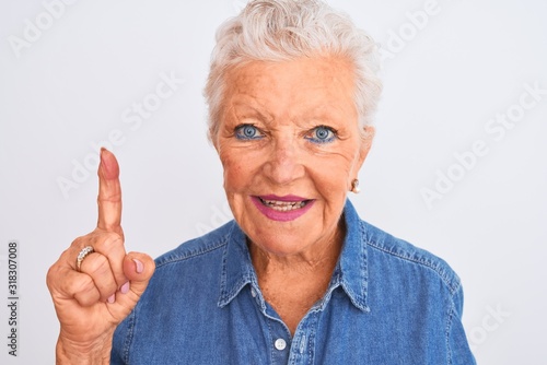 Senior grey-haired woman wearing casual denim shirt standing over isolated white background surprised with an idea or question pointing finger with happy face, number one