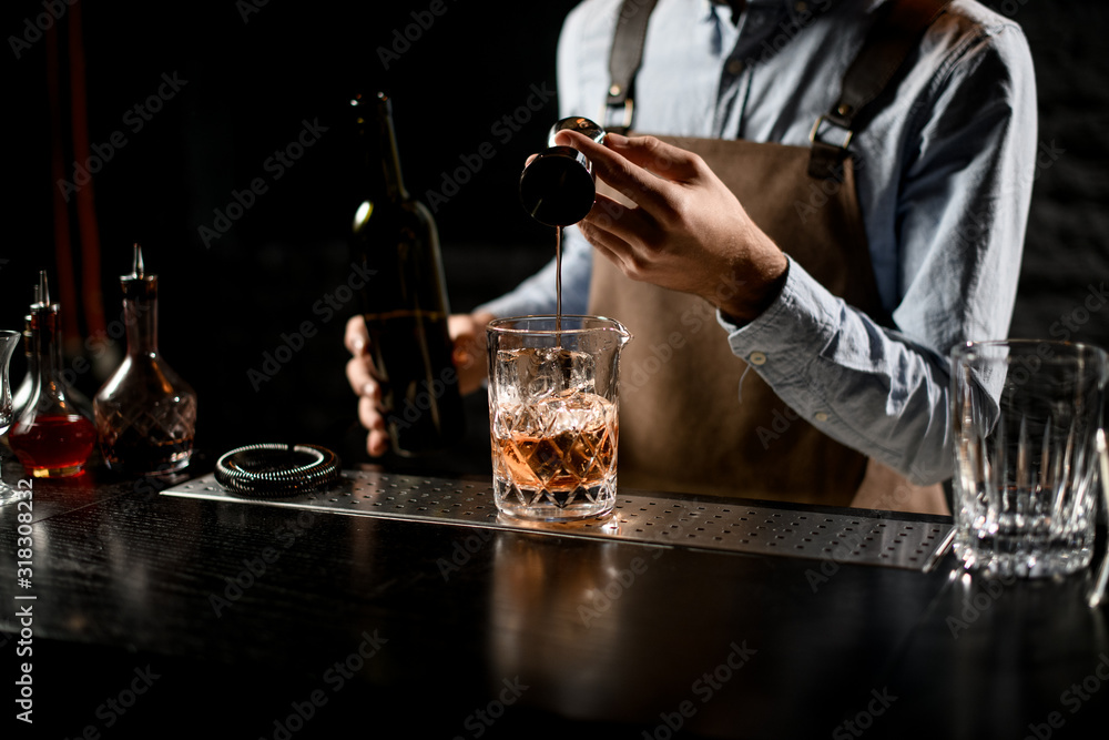Professional bartender pouring a golden alcoholic drink from the steel jigger to a glass
