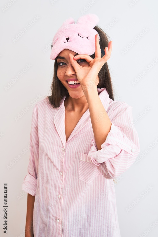 Young beautiful woman wearing sleep mask and pajama over isolated white background with happy face smiling doing ok sign with hand on eye looking through fingers Young beautiful woman wearing sleep mask and pajama over isolated white background with happy face smiling doing ok sign with hand on eye looking through fingers