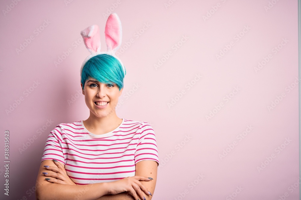 Young woman with fashion blue hair wearing easter rabbit ears over pink background happy face smiling with crossed arms looking at the camera. Positive person. Young woman with fashion blue hair wearing easter rabbit ears over pink background happy face smiling with crossed arms looking at the camera. Positive person.