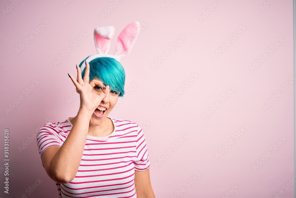 Young woman with fashion blue hair wearing easter rabbit ears over pink background doing ok gesture with hand smiling, eye looking through fingers with happy face.