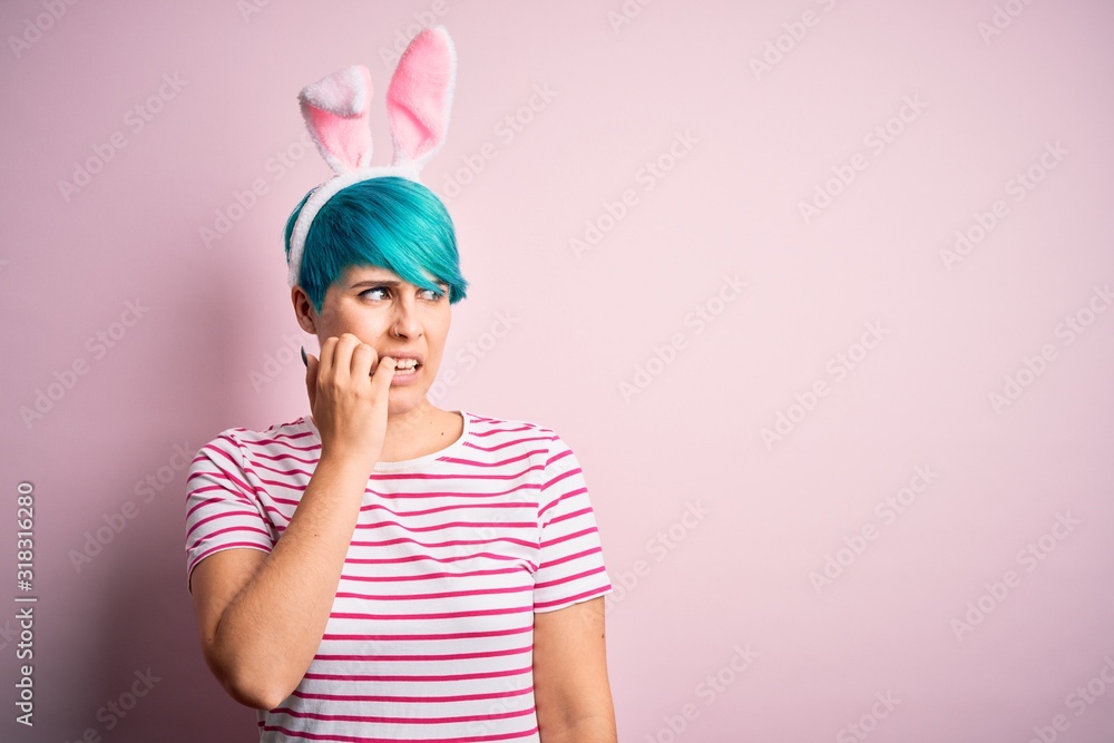 Young woman with fashion blue hair wearing easter rabbit ears over pink background looking stressed and nervous with hands on mouth biting nails. Anxiety problem. Young woman with fashion blue hair wearing easter rabbit ears over pink background looking stressed and nervous with hands on mouth biting nails. Anxiety problem.