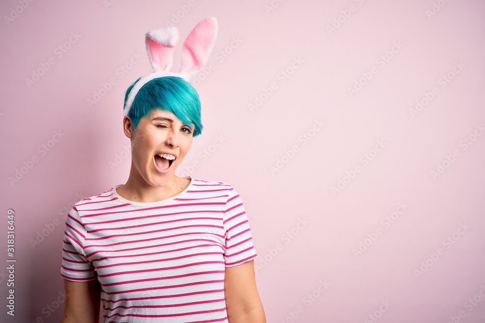 Young woman with fashion blue hair wearing easter rabbit ears over pink background winking looking at the camera with sexy expression, cheerful and happy face. Young woman with fashion blue hair wearing easter rabbit ears over pink background winking looking at the camera with sexy expression, cheerful and happy face.