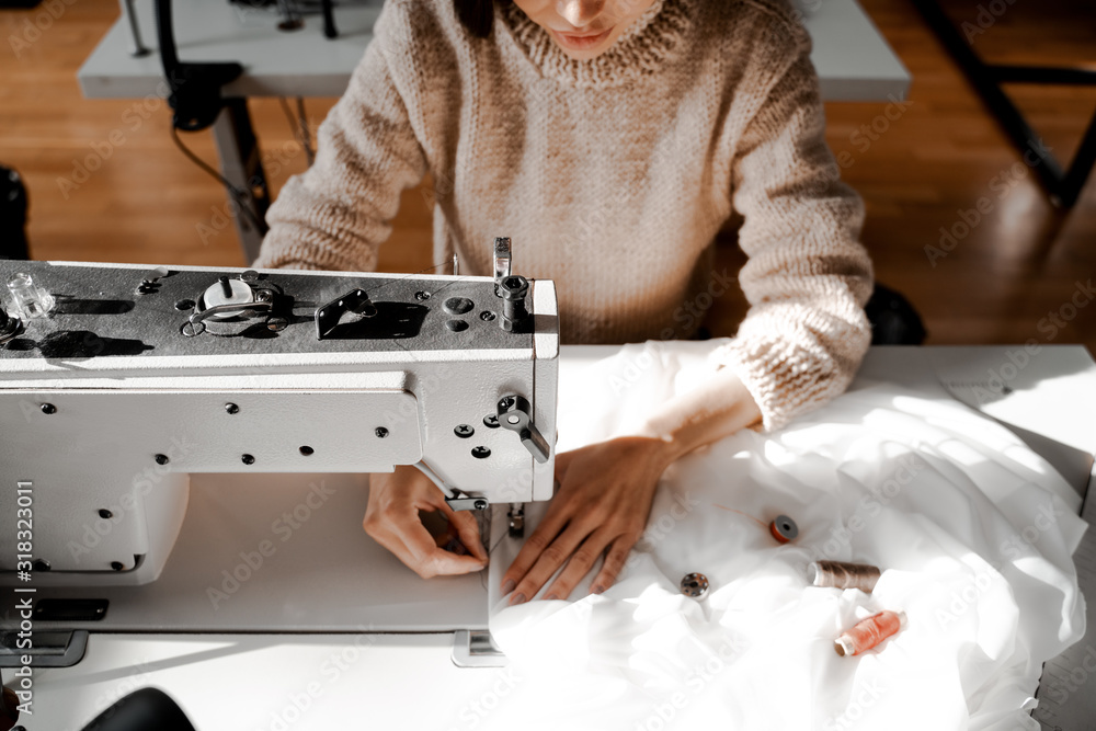 Woman seamstress at the sewing machine focused on sewing. Sewing, design work, tailoring studio, tailor, designer clothes, manufactory, in the process of creative development - obrazy, fototapety, plakaty 