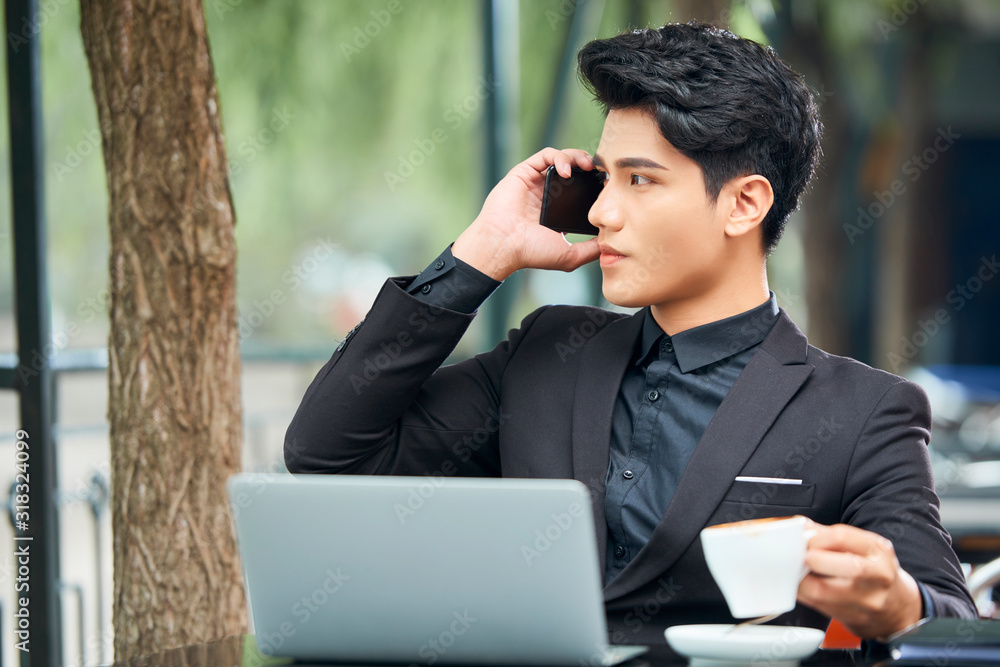 Modern Asian man sitting outdoors at cafe table with netbook on it talking on smartphone looking away Modern Asian man sitting outdoors at cafe table with netbook on it talking on smartphone looking away