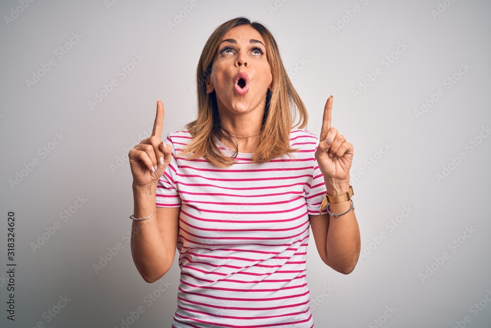 Middle age beautiful woman wearing casual striped t-shirt over isolated white background amazed and surprised looking up and pointing with fingers and raised arms. Middle age beautiful woman wearing casual striped t-shirt over isolated white background amazed and surprised looking up and pointing with fingers and raised arms.