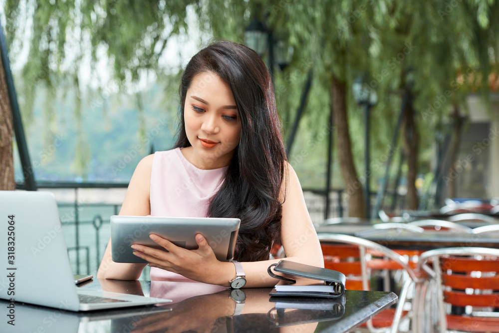 Horizontal portrait of beautiful Asian woman wearing pale pink dress sitting at cafe table using her tablet PC Horizontal portrait of beautiful Asian woman wearing pale pink dress sitting at cafe table using her tablet PC