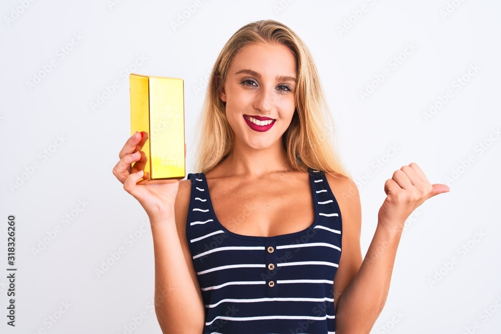 Young beautiful blond woman holding gold ingot standing over isolated white background pointing and showing with thumb up to the side with happy face smiling Young beautiful blond woman holding gold ingot standing over isolated white background pointing and showing with thumb up to the side with happy face smiling