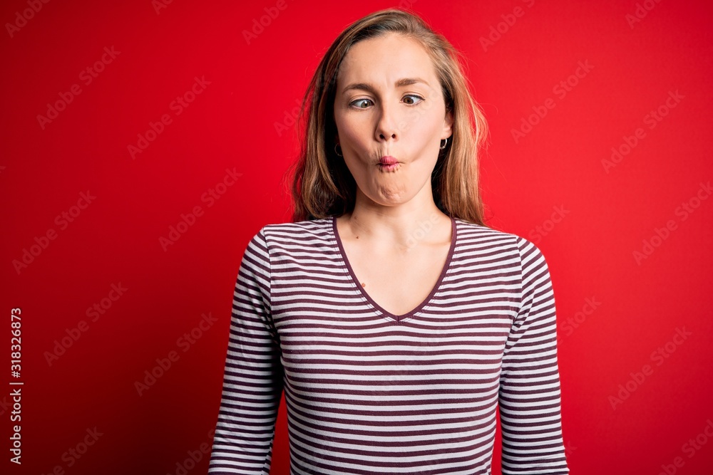 Young beautiful blonde woman wearing casual striped t-shirt over isolated red background making fish face with lips, crazy and comical gesture. Funny expression. Young beautiful blonde woman wearing casual striped t-shirt over isolated red background making fish face with lips, crazy and comical gesture. Funny expression.