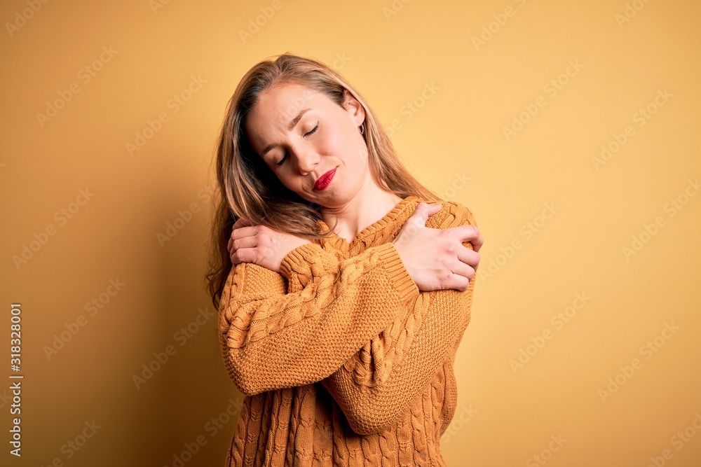 Young beautiful blonde woman wearing casual sweater standing over yellow background Hugging oneself happy and positive, smiling confident. Self love and self care Young beautiful blonde woman wearing casual sweater standing over yellow background Hugging oneself happy and positive, smiling confident. Self love and self care
