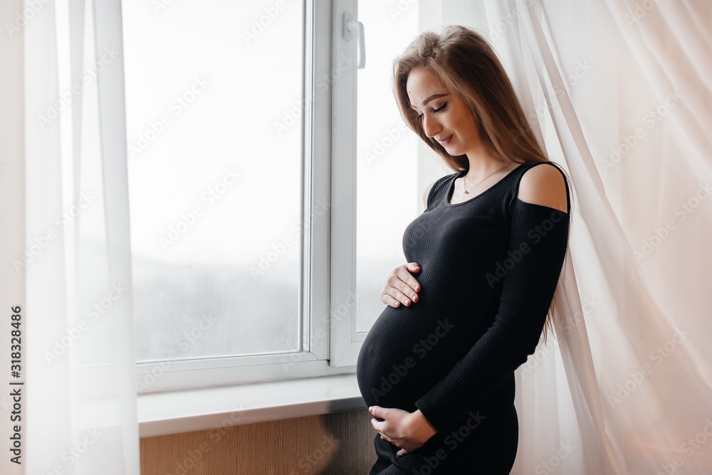 A beautiful pregnant girl is standing in a room near the window A beautiful pregnant girl is standing in a room near the window