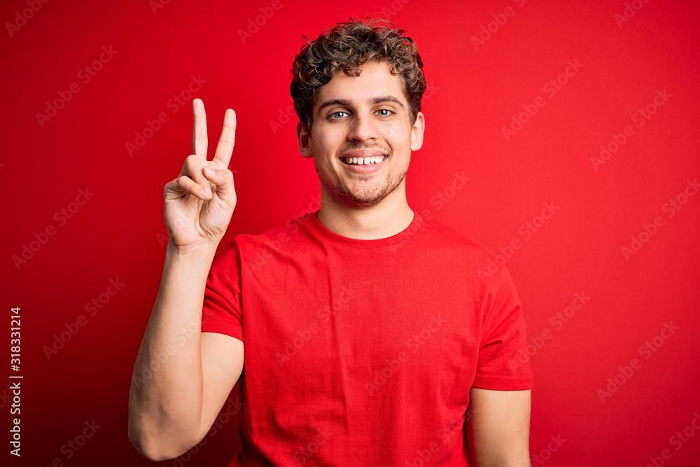 Naklejka premium Young blond handsome man with curly hair wearing casual t-shirt over red background showing and pointing up with fingers number two while smiling confident and happy.