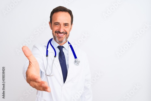 Middle age doctor man wearing coat and stethoscope standing over isolated white background smiling friendly offering handshake as greeting and welcoming. Successful business.