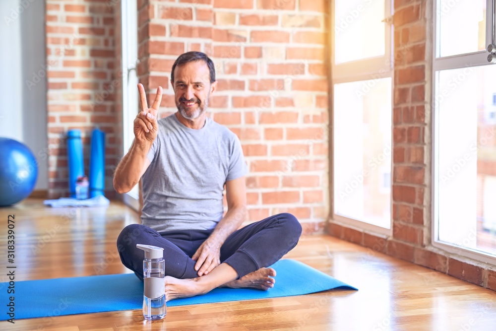 Middle age handsome sportman sitting on mat doing stretching yoga exercise at gym smiling looking to the camera showing fingers doing victory sign. Number two. Middle age handsome sportman sitting on mat doing stretching yoga exercise at gym smiling looking to the camera showing fingers doing victory sign. Number two.