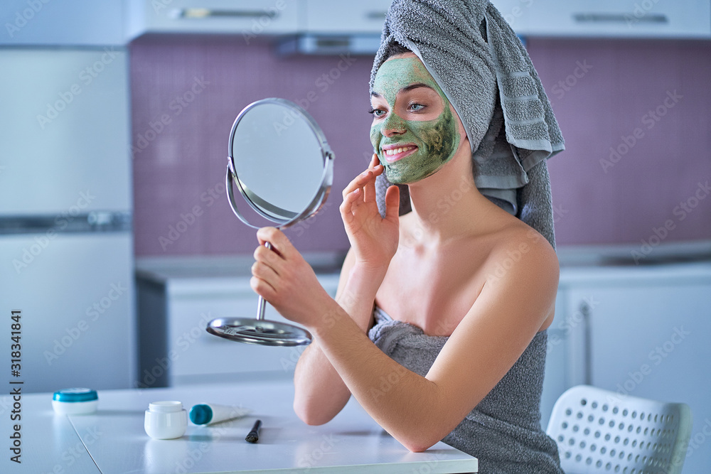 Φωτογραφία Smiling young happy healthy female in bath towel with green cleansing face clay mask and small round table mirror during spa day at home