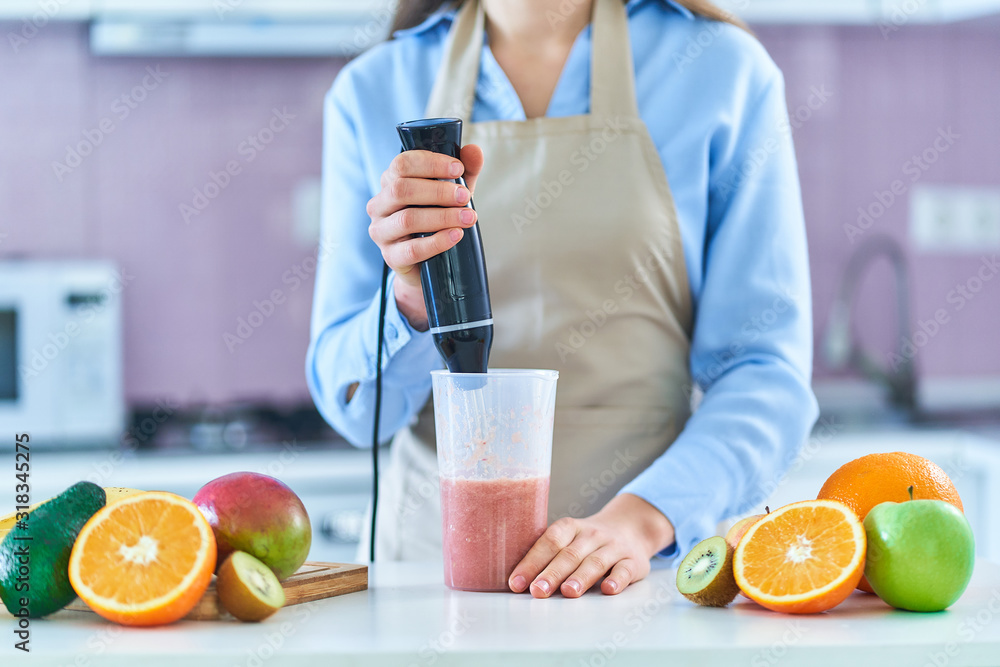 Woman in apron uses hand blender to mixing fresh fruits for prepare smoothie in the kitchen at home Woman in apron uses hand blender to mixing fresh fruits for prepare smoothie in the kitchen at home