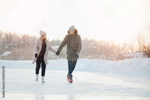 Girl and man are holding hands looking each other and eating on frozen ice of lake on skates. In background is sunset