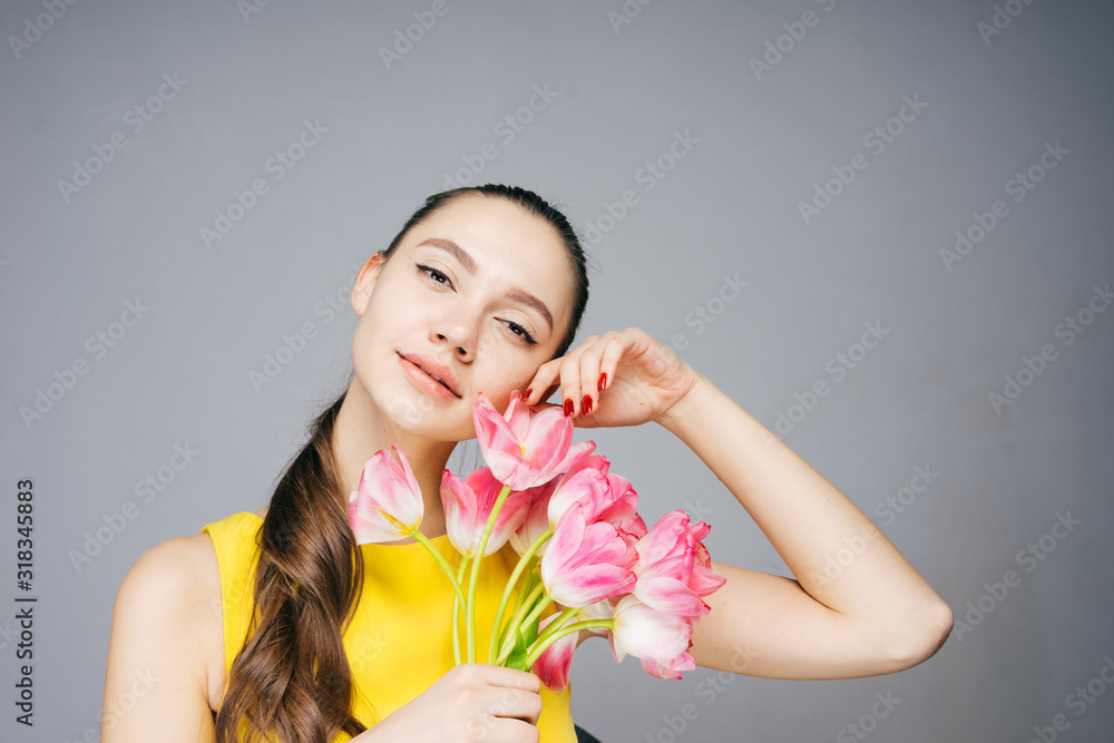 a beautiful girl in a bright yellow dress with tenderness leaned a bouquet of tulips against her cheek. gray background