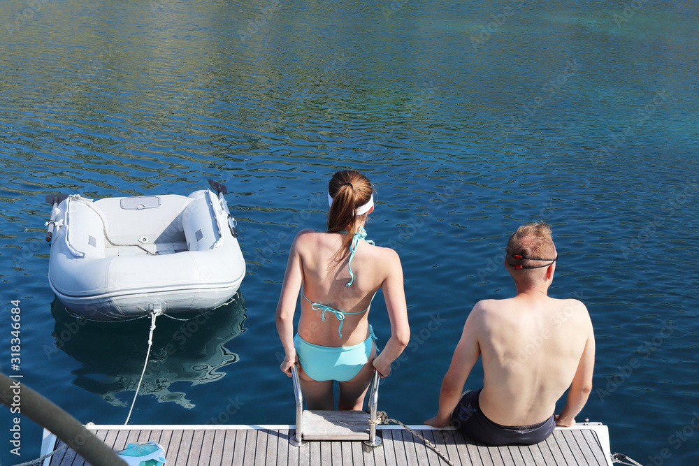 A young couple a guy and a girl in a blue swimsuit are standing on the yacht ramp and are going to take a dip in the sea. Exclusive holiday at the sea. Relax in a paradise bay. Dinghi moored