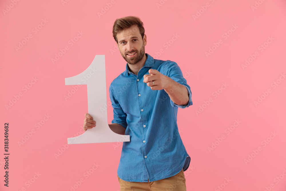 happy casual guy holding number one sign and pointing finger - obrazy, fototapety, plakaty 