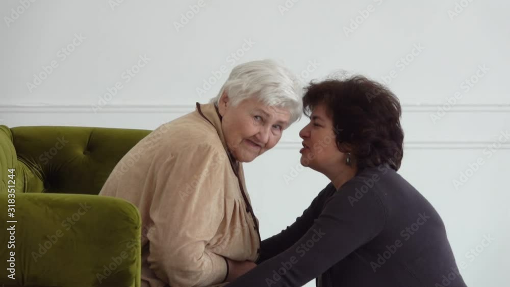 Beautiful middle-aged woman tells a joke to her elderly mother sitting on a sofa in a white room. Women are laughing loudly Beautiful middle-aged woman tells a joke to her elderly mother sitting on a sofa in a white room. Women are laughing loudly